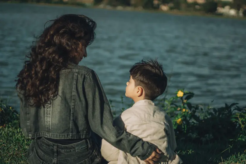A mother with her arm around her young son, sitting by a lake