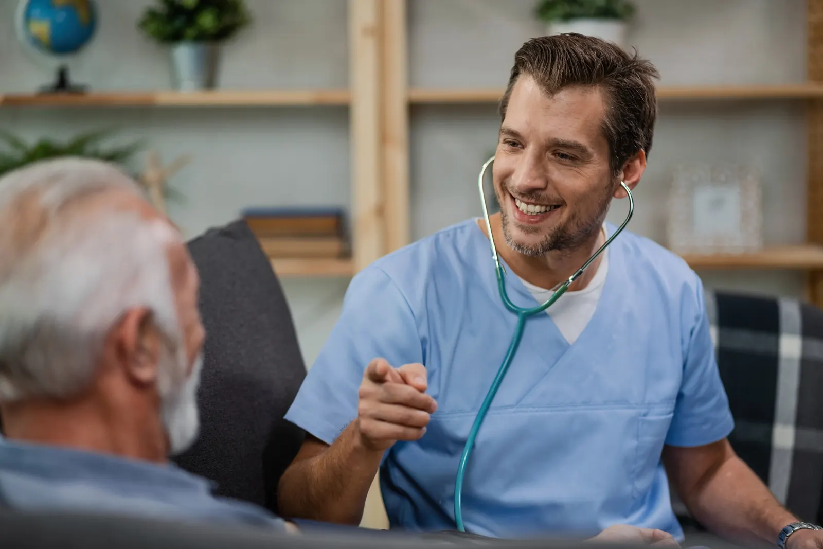 A smiling doctor in blue scrubs talking with an older patient during a home visit