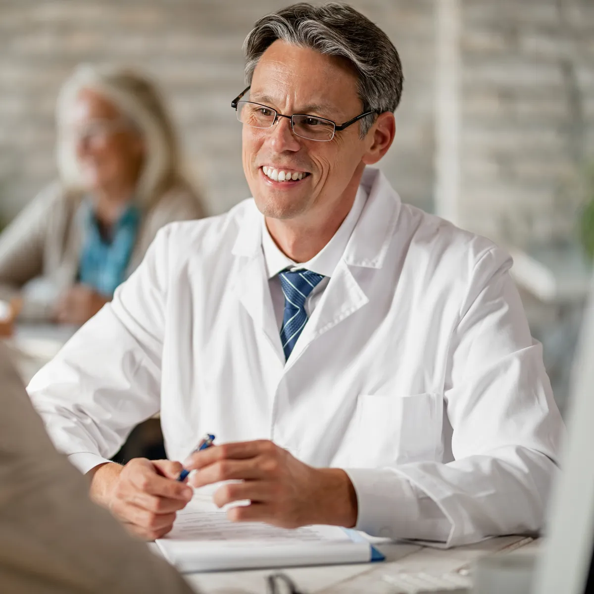 A smiling doctor in a white coat talking with an older patient across a desk