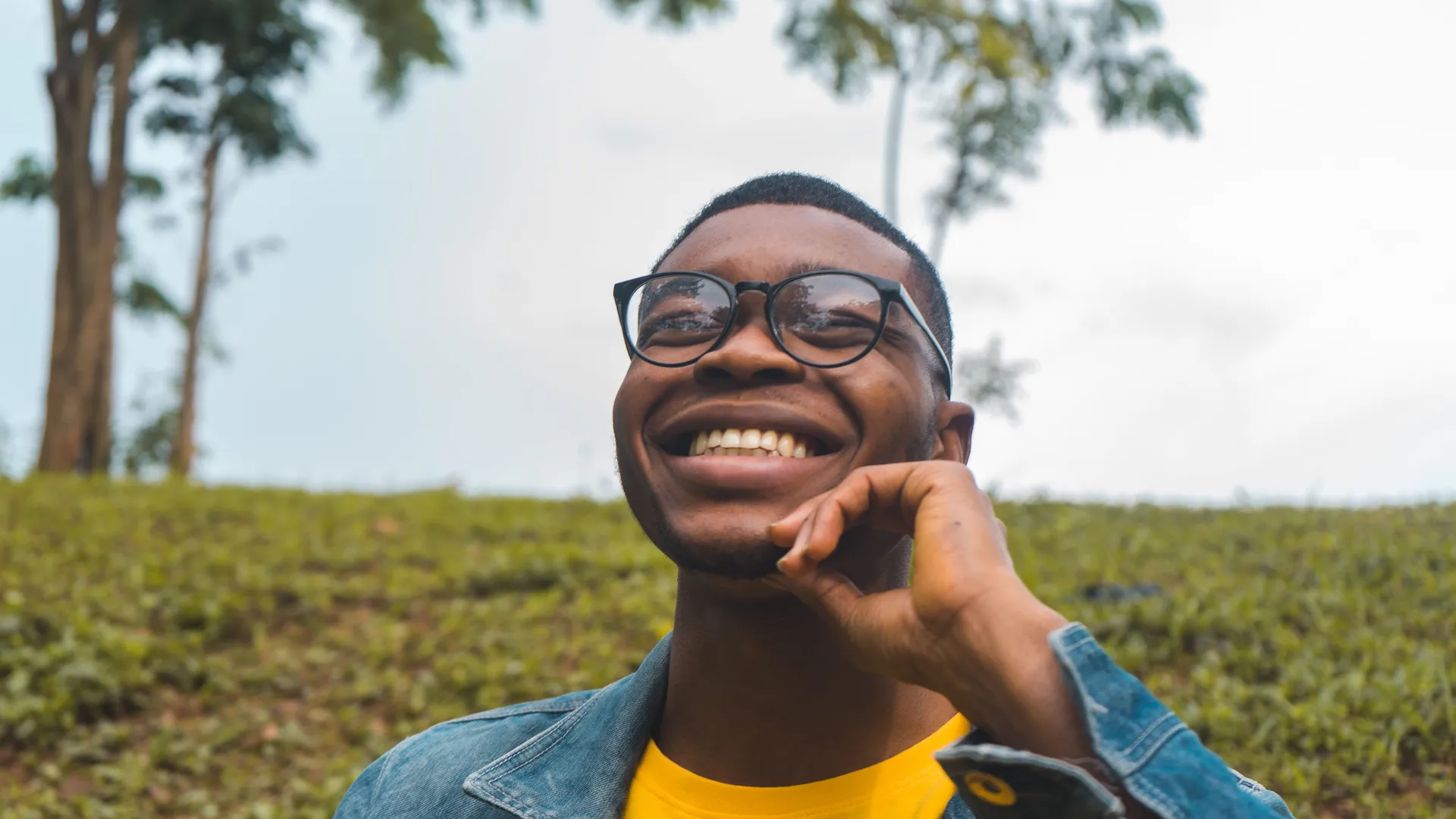 James laughing outdoors in a denim jacket and glasses